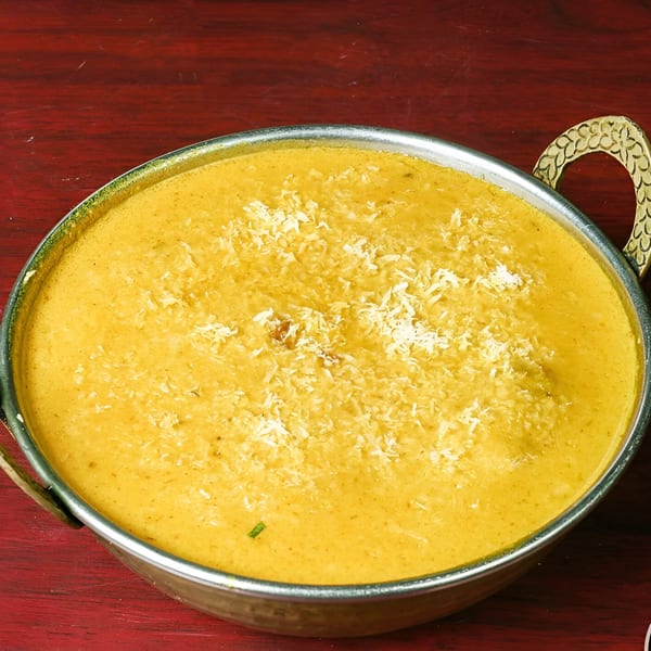 A metal bowl of creamy yellow Indian curry, garnished with shredded coconut, on a red wooden surface.