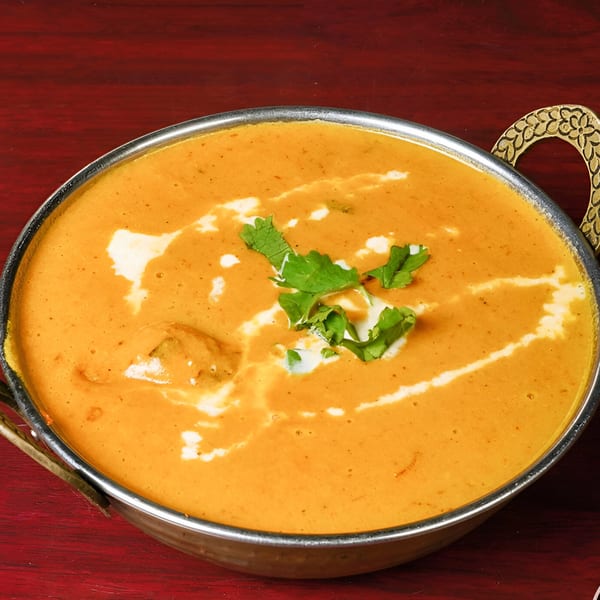 A close-up of creamy orange Indian curry with white swirls and green garnish in a metal bowl.
